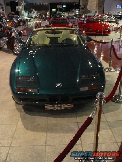 Green convertible sports car with a Union Jack plate in front, parked indoors with other vehicles and motorcycles