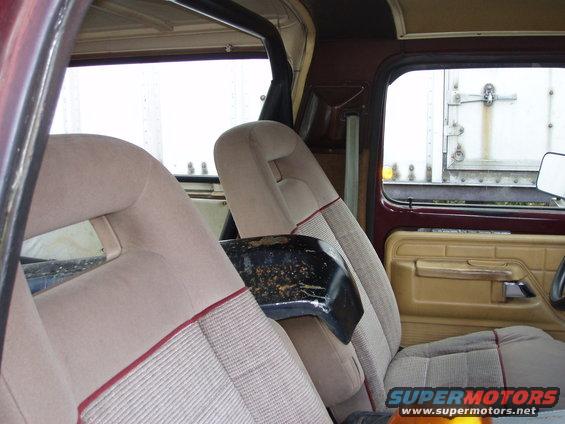 Interior shot of a vehicle, showing tan seats with red trim and a black center console piece. A trailer is visible through the window