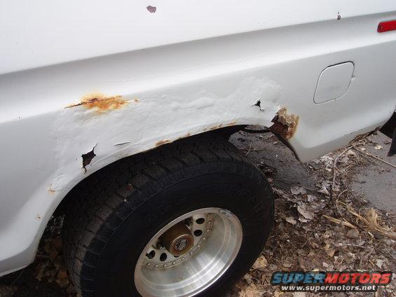 White vehicle's rear fender shows significant rust and holes. Tire and wheel are visible