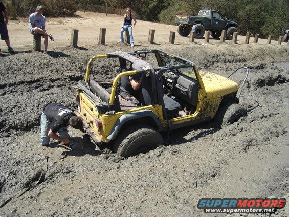 Yellow Jeep Wrangler stuck in mud, with a person trying to free it and a green pickup truck in the background