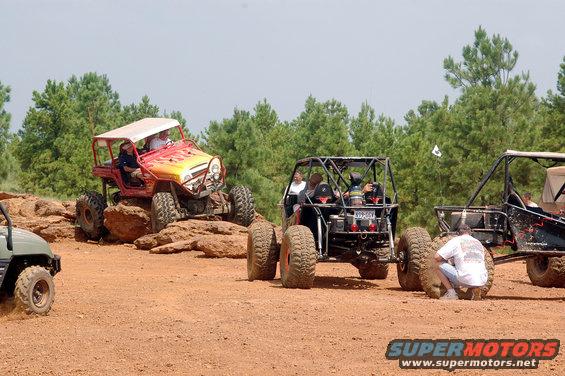 Several off-road vehicles, including a red Jeep and black buggies, navigate a rocky, dirt trail with trees in the background