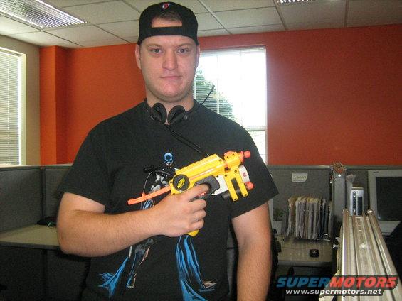 Guy in office wearing a baseball cap and headphones, holding a yellow toy gun