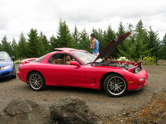Red Mazda RX-7 with hood open, parked next to a blue car on a gravel road with trees in the background