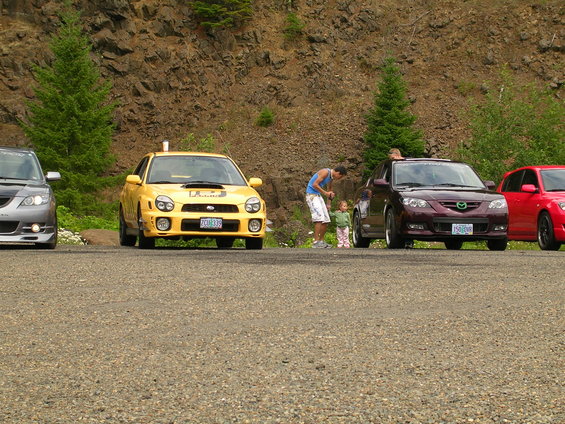Yellow Subaru WRX, a maroon Mazda 3, and a red car parked on gravel with a rocky hill behind them