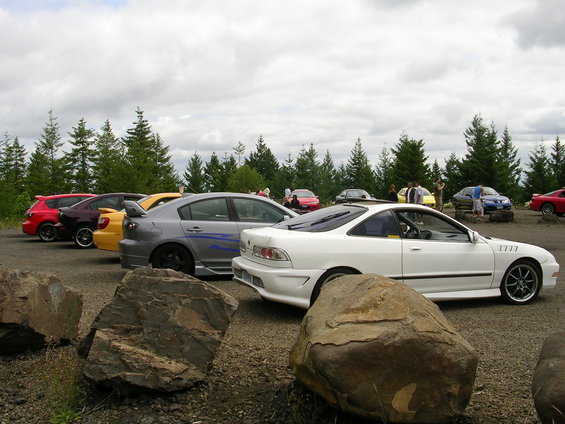 Several cars are parked in a lot with trees and a cloudy sky in the background. A white coupe is in the foreground