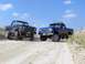 1987 Ford Bronco - Two lifted Ford pickup trucks are parked on a dusty dirt road under a blue sky.