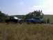 1987 Ford Bronco - Two pickup trucks are parked in a field with trees and hills in the background under a partly cloudy sky.
