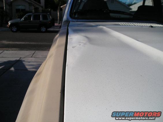 Close-up of a dented white truck hood, with another SUV parked on the street in the background
