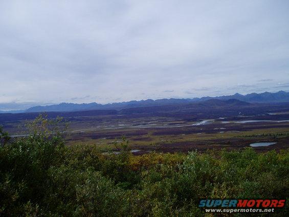 Mountain range in the distance with a winding river and marshy ground in the foreground, under a cloudy sky