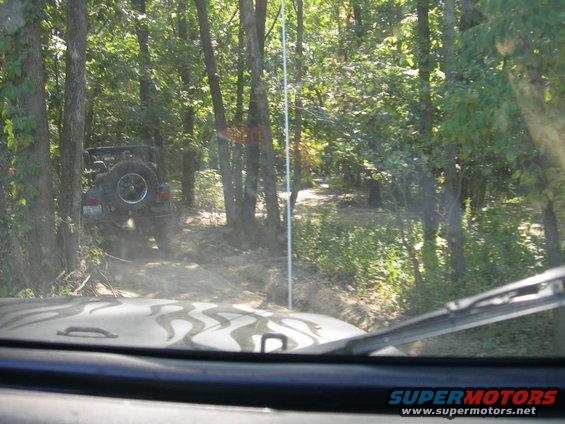 Two Jeep Wranglers driving on a dirt path through a forest. One is in front, seen from the dashboard, and the other is further ahead