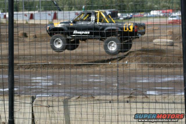 Black pickup truck with yellow roll cage jumping over dirt track. Has 'Snap-on' text on the door and a yellow 'K' on the tailgate