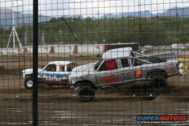 Two modified pickup trucks racing on a dirt track. One has 'DUSTER' on its side, the other has 'JAMMIN SALMON'