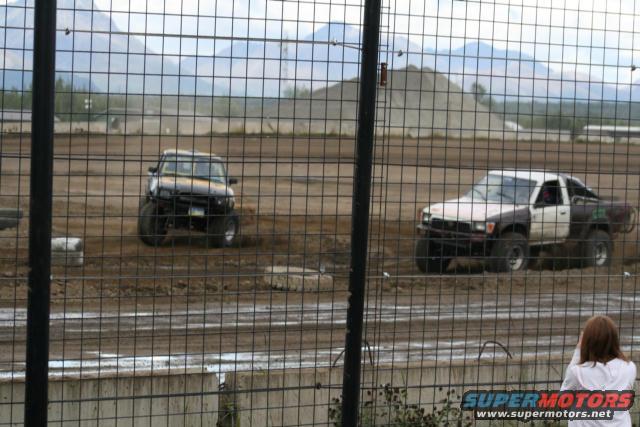 Two modified trucks on a dirt track, one black and one white and brown, with large tires and lifted suspension