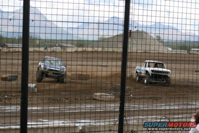 Two off-road trucks race on a dirt track with mountains in the background