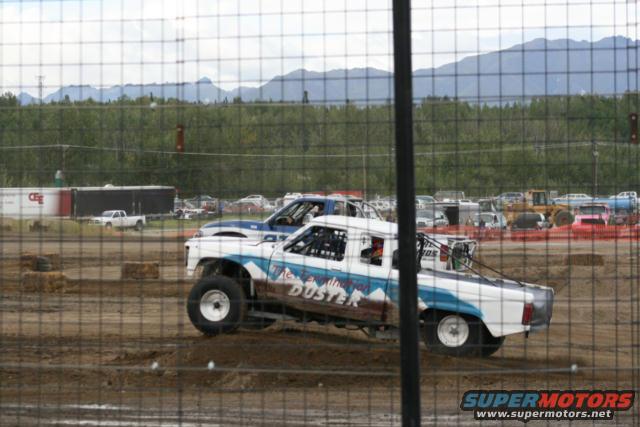 Modified pickup truck with "DUSTER" graphics racing on a dirt track, another blue truck behind it. Mountains in background