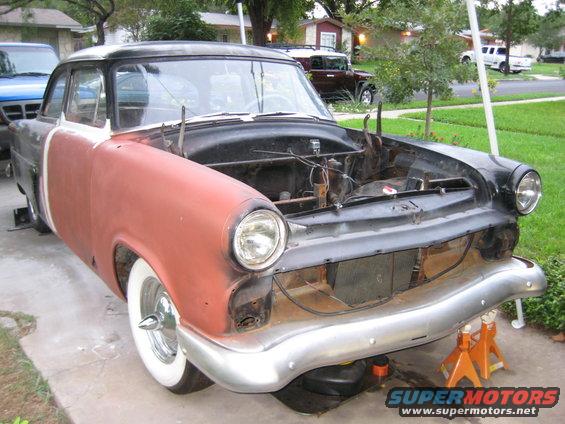 Front end of a 1954 Ford sedan with hood removed, sitting on jack stands. Red primer on front fender