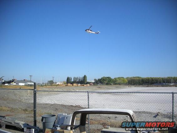 Helicopter lifting an object over a construction site with a partial vehicle frame in the foreground