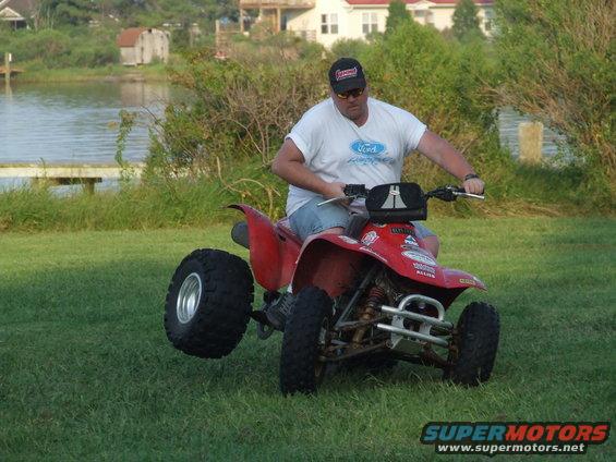Man on red ATV with one wheel up, grassy area, water and houses in background