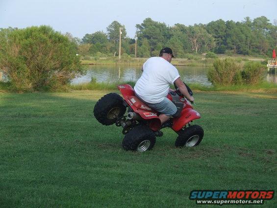 Red ATV with a man riding it on its rear wheels in a grassy field near a body of water