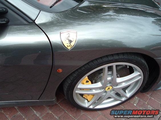 Gray Ferrari fender with a yellow and black prancing horse emblem and a silver multi-spoke wheel with yellow brake calipers