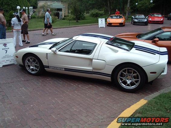 White Ford GT with blue stripes parked on brick pavement, other sports cars in background