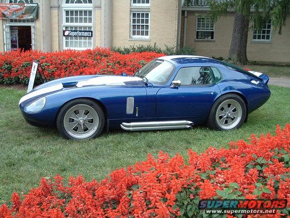 Blue Shelby Cobra Daytona Coupe replica with white stripes parked on grass in front of a building with red flowers