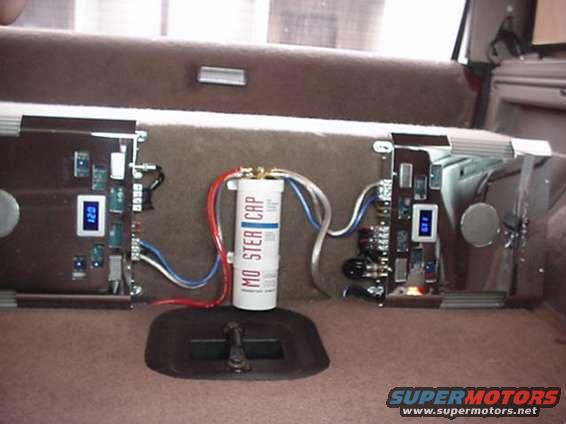 Car audio capacitor labeled 'MO STER CAP' between two amplifiers with blue digital displays