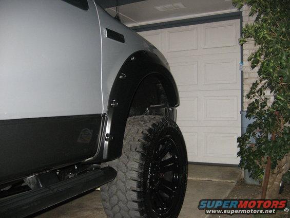 Side view of a truck's front fender and large off-road tire with black rims. Garage door in background