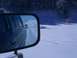 1979 Ford Bronco - A green SUV is seen from its side mirror driving on a snow-covered road with another vehicle ahead.