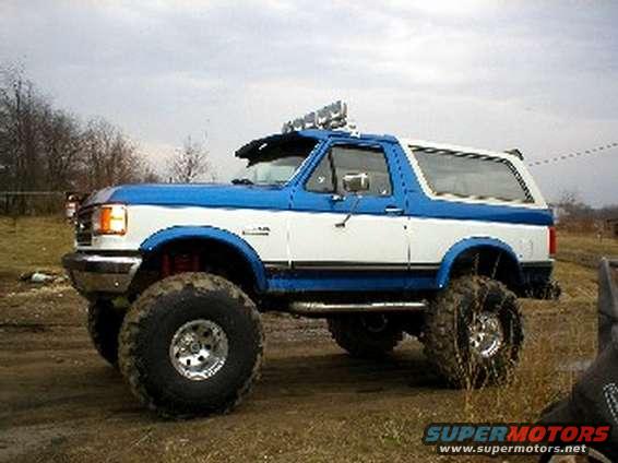 Blue and white lifted Ford Bronco with large off-road tires, aftermarket light bar, and side exhaust