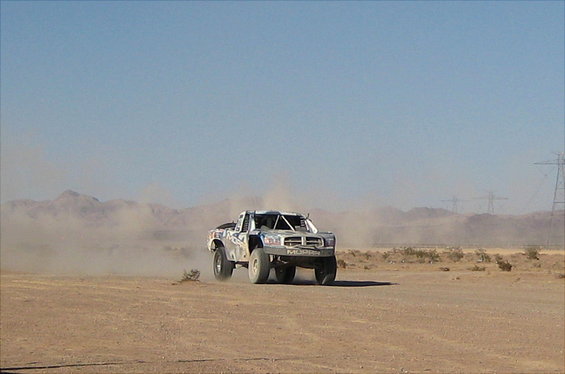 Off-road truck kicking up dust on a dirt track with mountains and power lines in the background