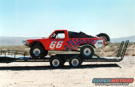 Red off-road truck with number 66 and checkered flag graphics on a trailer in a desert landscape