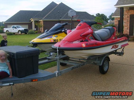 Two jet skis, one red and one yellow, on a trailer in front of a house. A white pickup truck is parked in the driveway