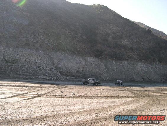 Two off-road vehicles driving on a dry, sandy riverbed with tire tracks, a large rocky hill in the background