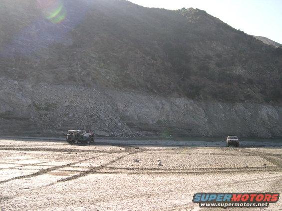 Two SUVs driving on a dry, dusty ground with tire tracks, near a rocky hillside