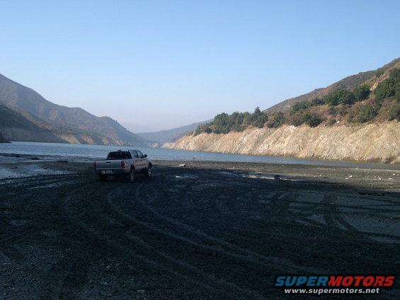 Pickup truck parked on a dirt area next to a large body of water with mountains in the background