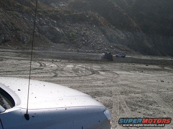 Silver SUV hood in foreground, two SUVs and bulldozer in background on dusty terrain