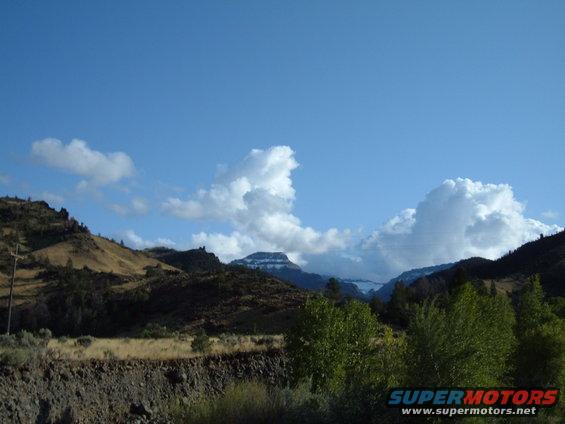 Mountain range under a blue sky with fluffy white clouds. Dry hills in foreground with green trees