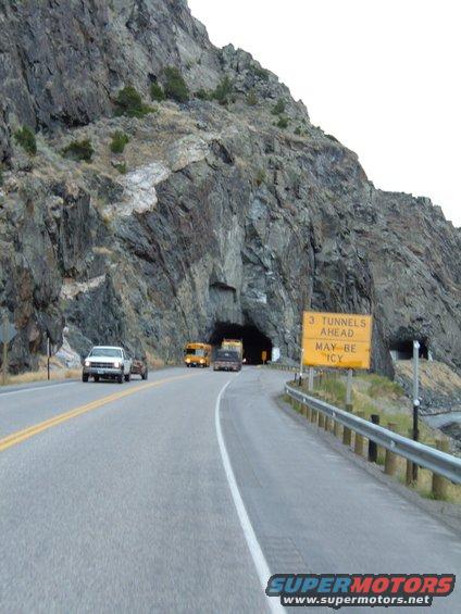 Road leading to a tunnel entrance in a rocky mountainside, with a white pickup truck, a yellow school bus, and other vehicles visible