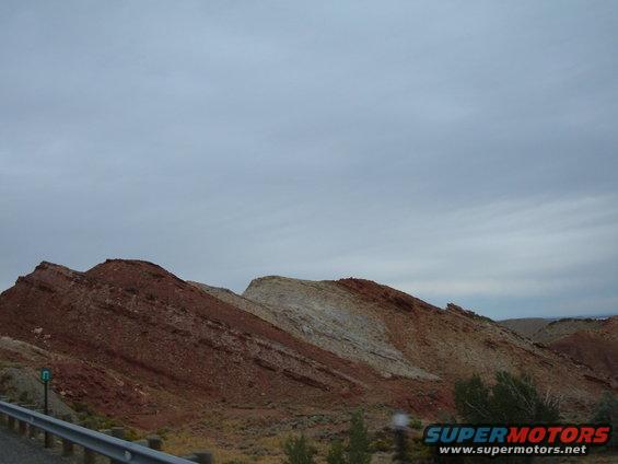 Red and white rock formations under a cloudy sky. Guardrail in foreground