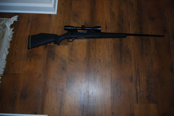 Black rifle with a scope mounted on top, lying on a wooden floor