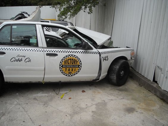 White taxi cab with front end damage, hood crumpled, windshield cracked. "Victory Metropolitan Taxi" logo on door