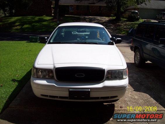 White 2003 Ford Crown Victoria parked in driveway, front view. Black grille, headlights visible. Another SUV parked to the right