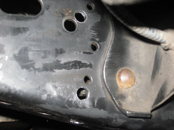 Close-up of a metal vehicle frame component with multiple drilled holes and a rusty bolt head