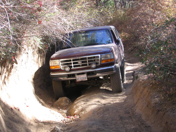 Maroon Ford Bronco navigating a narrow, rutted dirt trail with steep sides