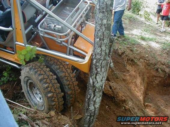 Off-road buggy with large tires stuck in dirt, leaning against a tree. People stand nearby
