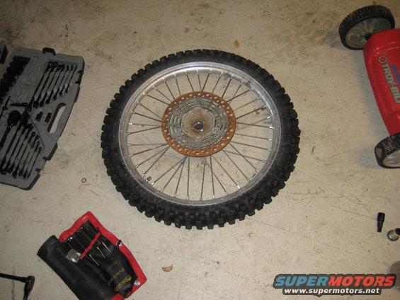 Motorcycle wheel with knobby tire and sprocket, next to tool kits and a red lawnmower