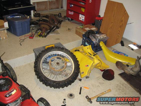 Yellow motorcycle fuel tank and seat with a large wheel and other parts scattered on a garage floor