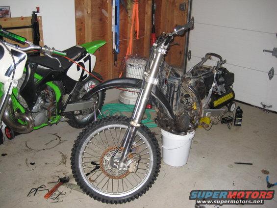 Motorcycle front wheel and engine assembly in a white bucket, with another green and white dirt bike to the left
