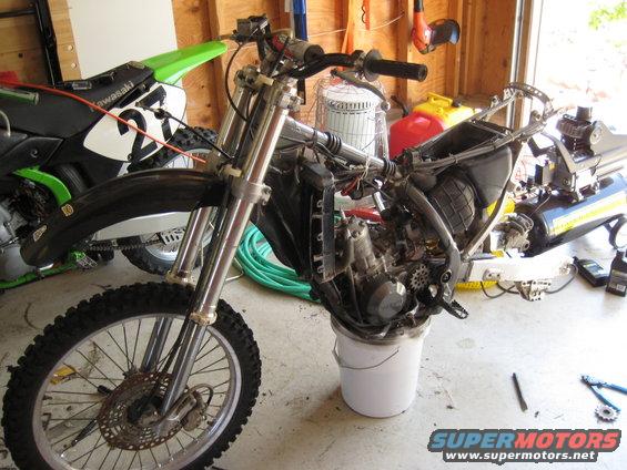 Motorcycle frame on a bucket, front wheel and engine visible. Another green motorcycle is behind it